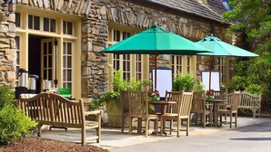 A stone building with a rustic exterior, outdoor seating with wooden chairs and tables, and a green umbrella providing shade. - Graylyn Estate (Winston-Salem)