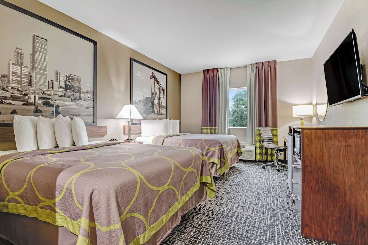 A hotel room with two beds, a desk, a TV, and a cityscape painting on the wall.