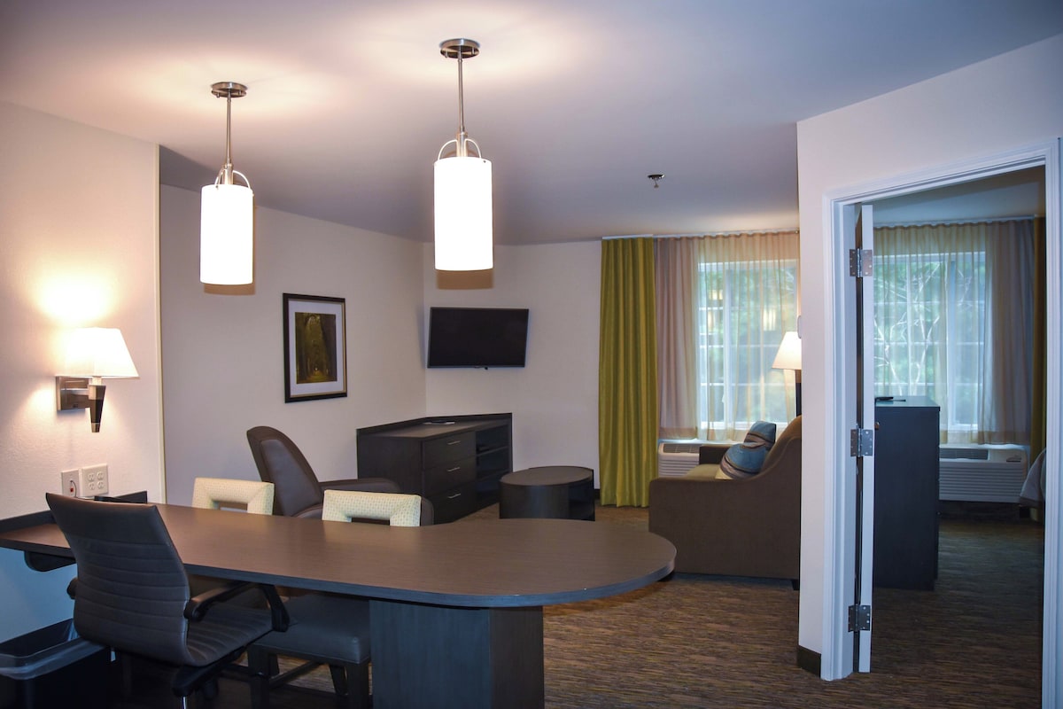 Een hotelkamer met een bureau, stoelen, een televisie, een flatscreen televisie aan de muur, een groot raam met gordijnen, en een deur naar een andere kamer.
