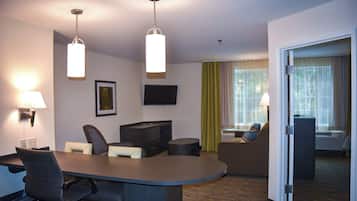 A hotel room with a desk, chairs, a television, a flat-screen TV mounted on the wall, a large window with curtains, and a door leading to another room.