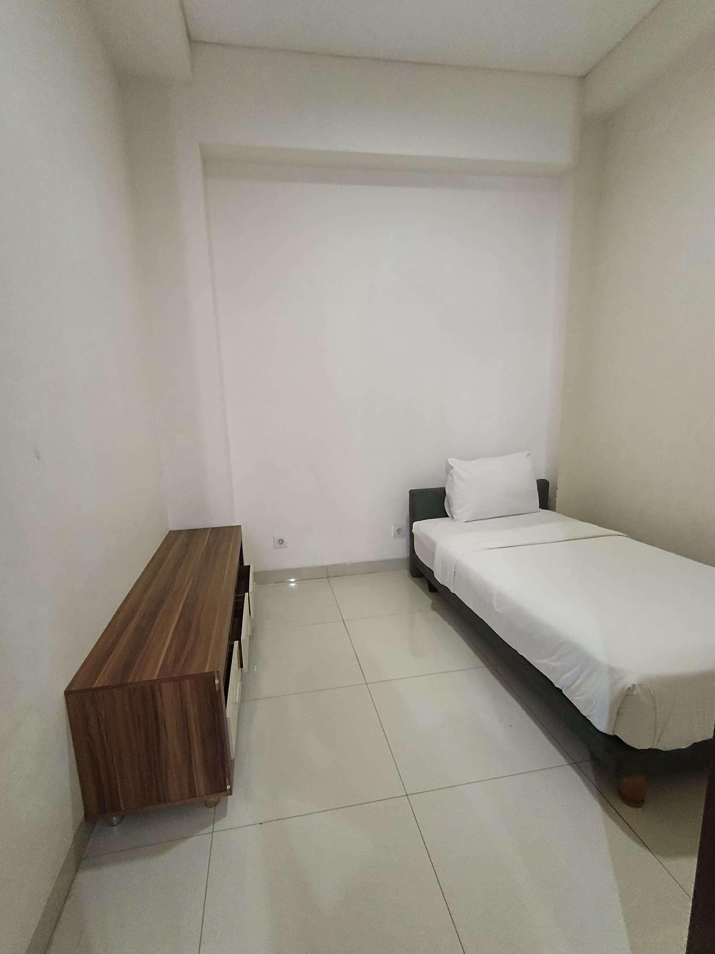 A small, clean room with a single bed, a wooden bench, and a tiled floor.