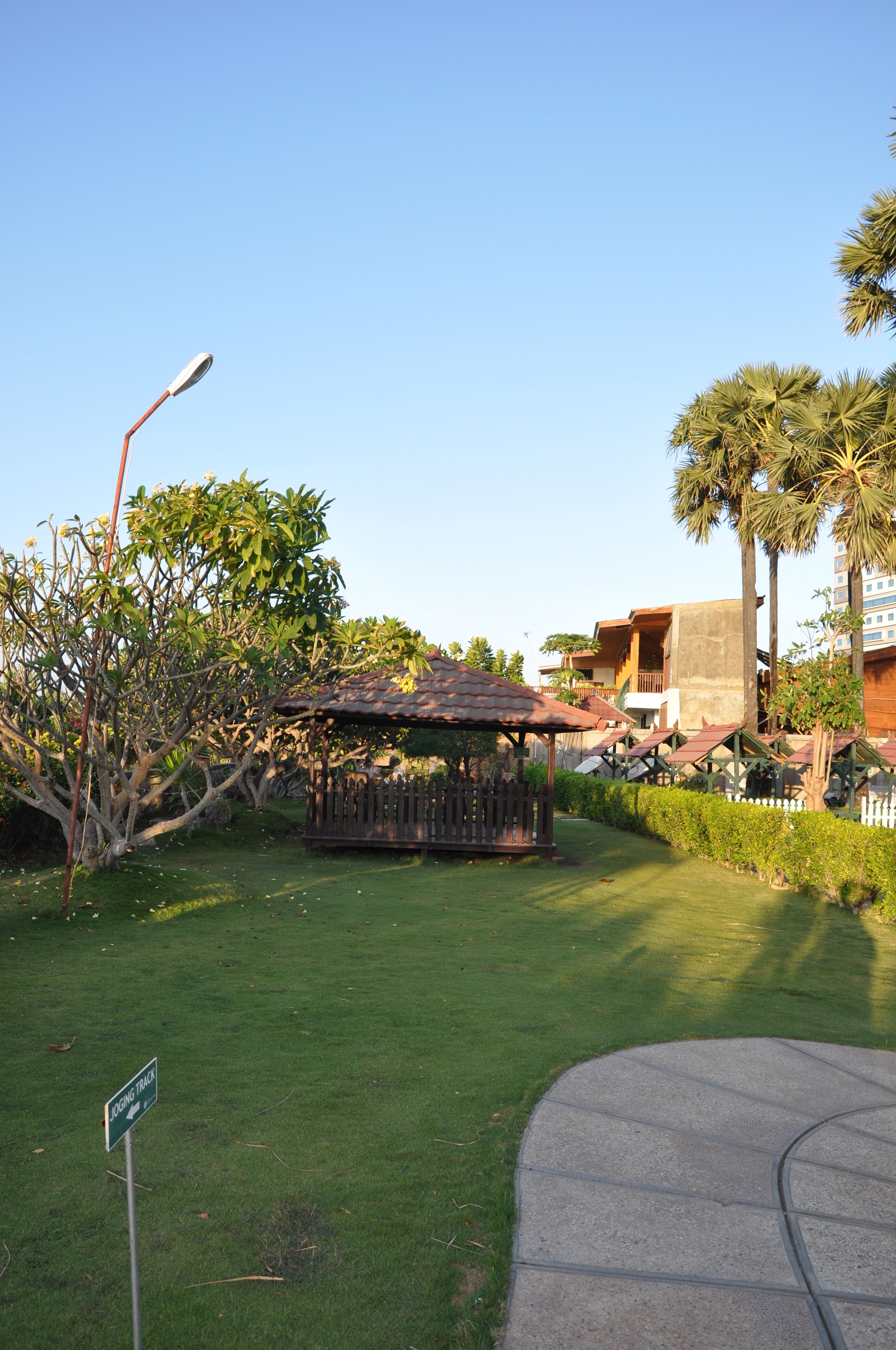 A gazebo in a well-maintained garden with a clear sky, surrounded by palm trees and a building with a flat roof in the background.