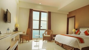 A hotel room with a bed, a desk, a chair, a small table, a TV, and a large window with curtains. - Hotel On The Rock (Kupang)