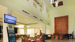 A hotel lobby with a reception desk, a television, wicker chairs, a grand piano, and a large wooden door. - Hotel On The Rock (Kupang)