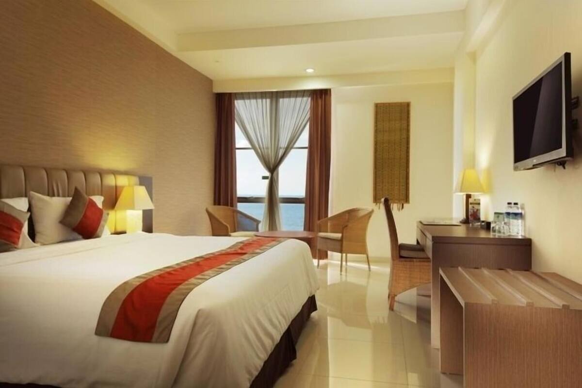A hotel room with a bed, a desk, a chair, a television, and a view of the sea.