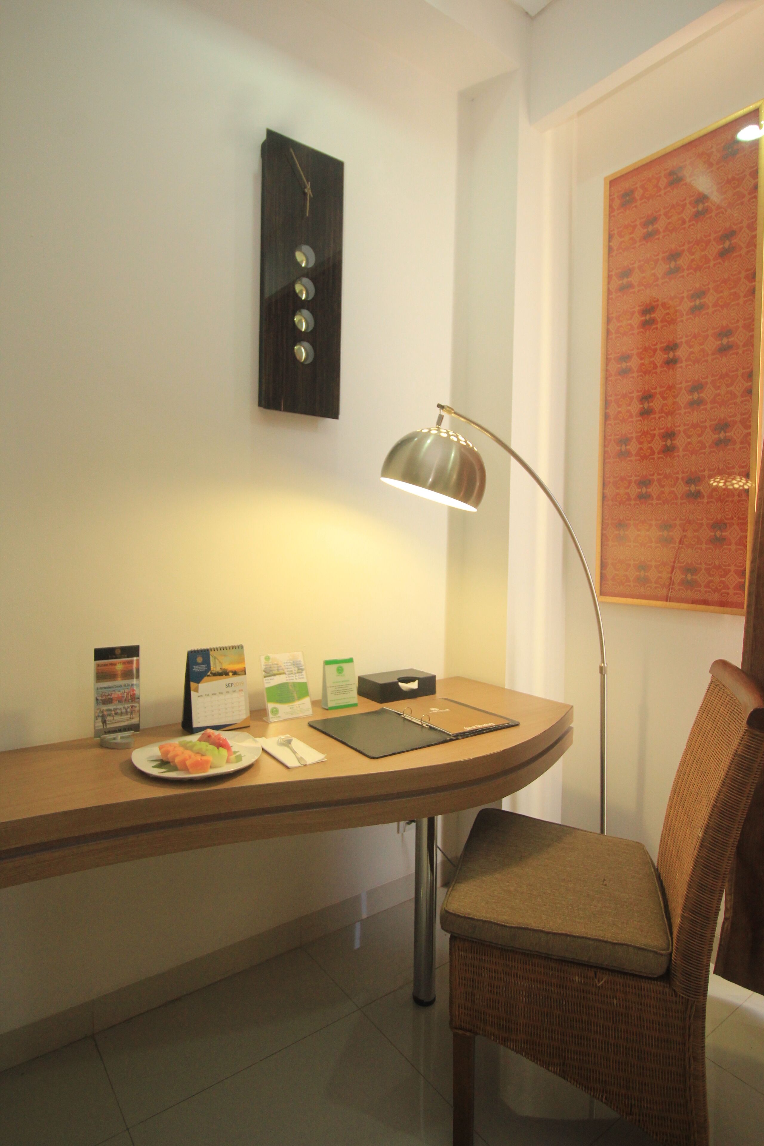 A hotel room with a desk, a chair, a clock, a lamp, and a framed picture on the wall.
