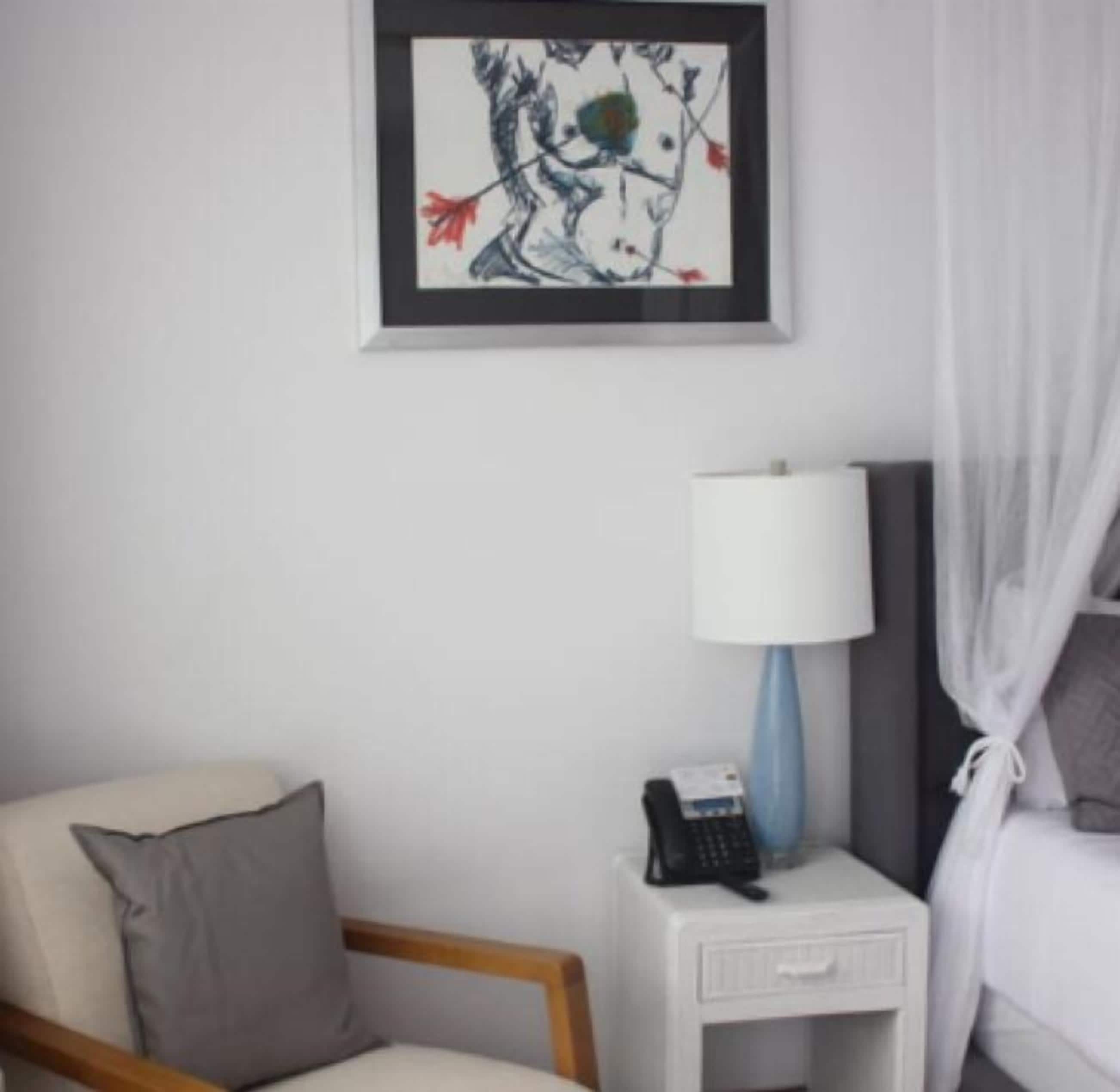 A hotel room with a bed, a bedside table with a lamp, a chair, and a framed artwork on the wall.