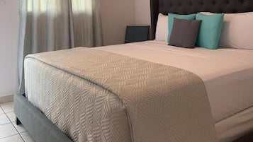 A neatly made bed with a patterned bedspread, a dark upholstered headboard, and a window with sheer curtains.