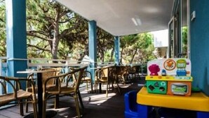 An outdoor seating area with wooden chairs and tables, a covered section with a blue railing, and a children's play area with colorful toys. - Hotel Mare Live (Jesolo)
