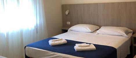 Una habitación de hotel con cama, mesitas de noche, un banco, una lámpara colgante y una obra de arte en la pared.