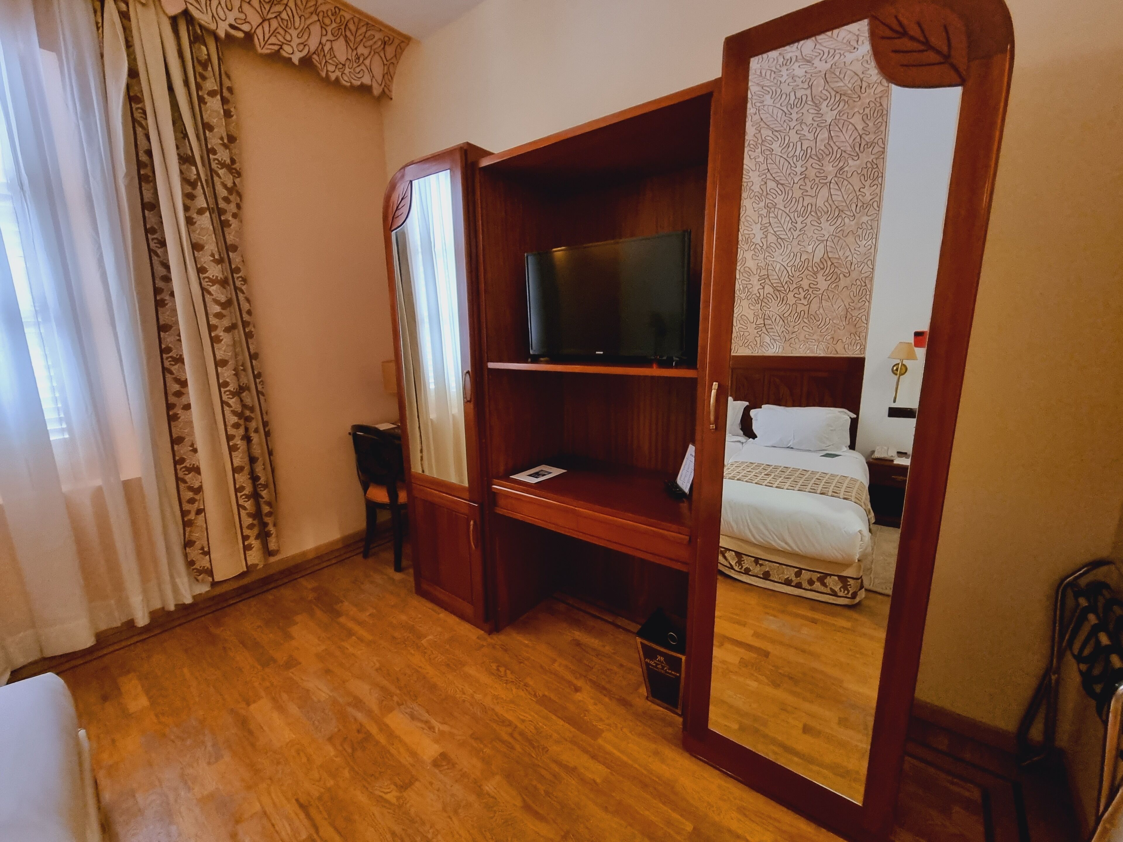 A hotel room with a wooden floor, a bed with a patterned headboard, a television on a wooden stand, a desk with a chair, and a mirror reflecting the room.