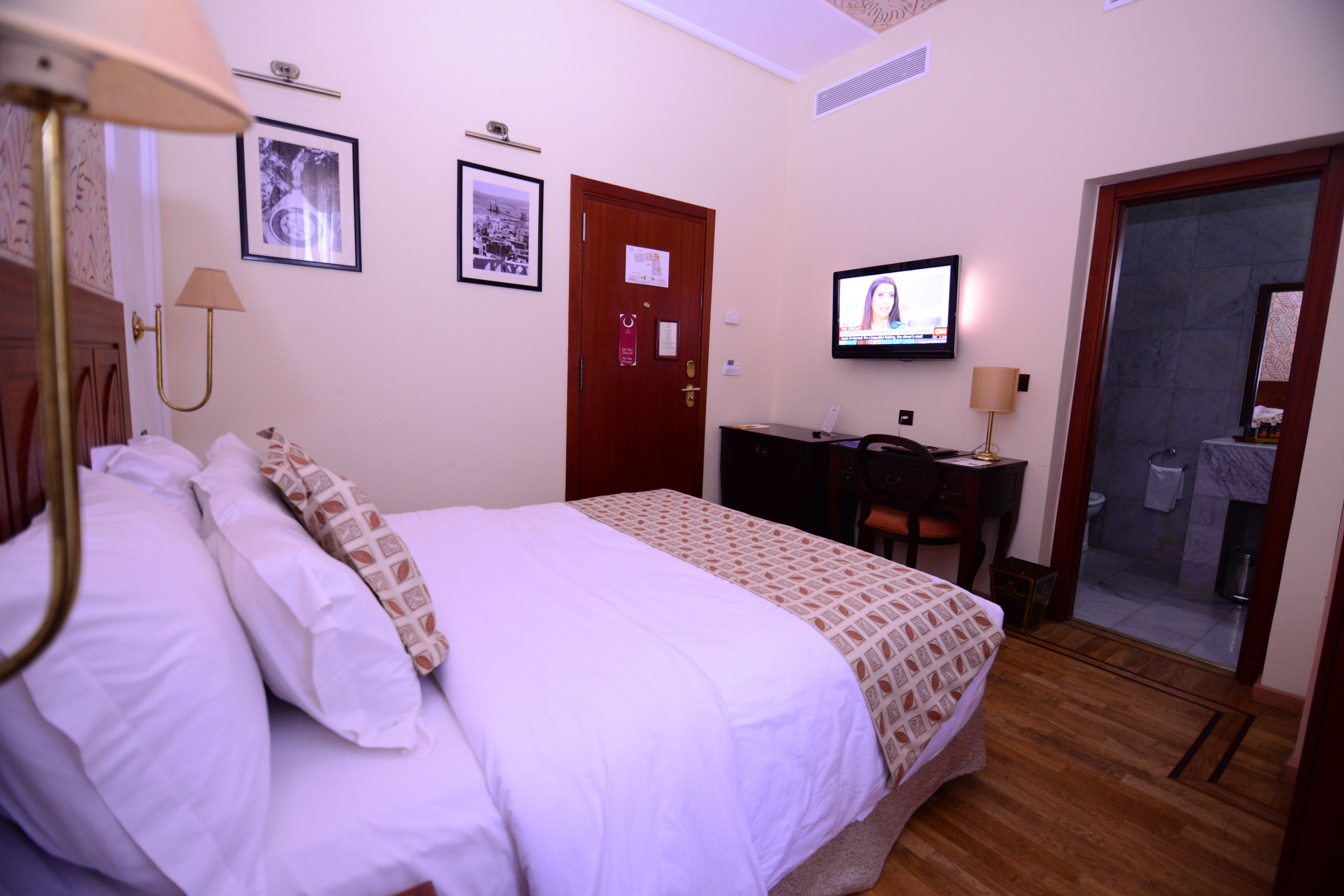Una camera d'albergo con un letto, una scrivania, una televisione e un bagno visibile attraverso una porta di vetro.