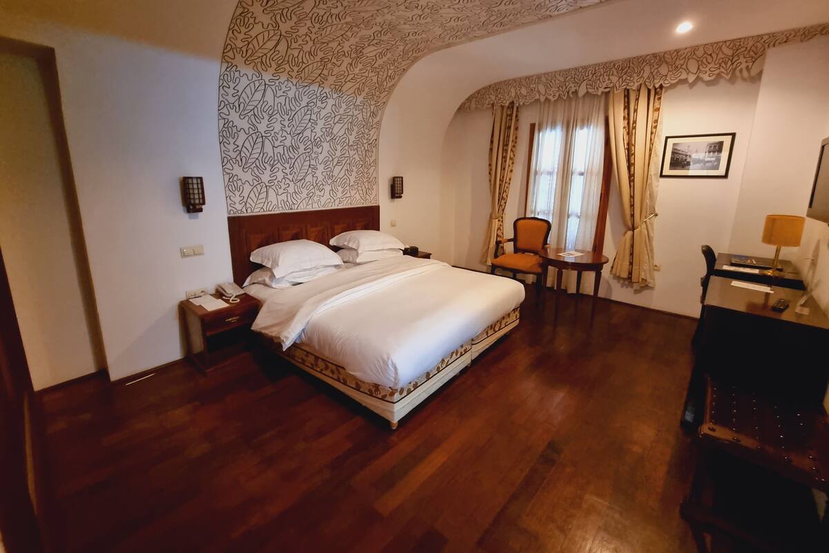 A hotel room with a large bed, a desk with a lamp, a chair, and a small table.