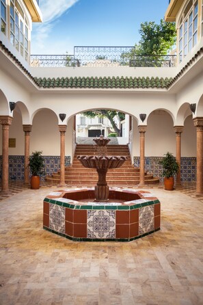 Une cour intérieure avec une fontaine centrale, un sol carrelé et des arcades.