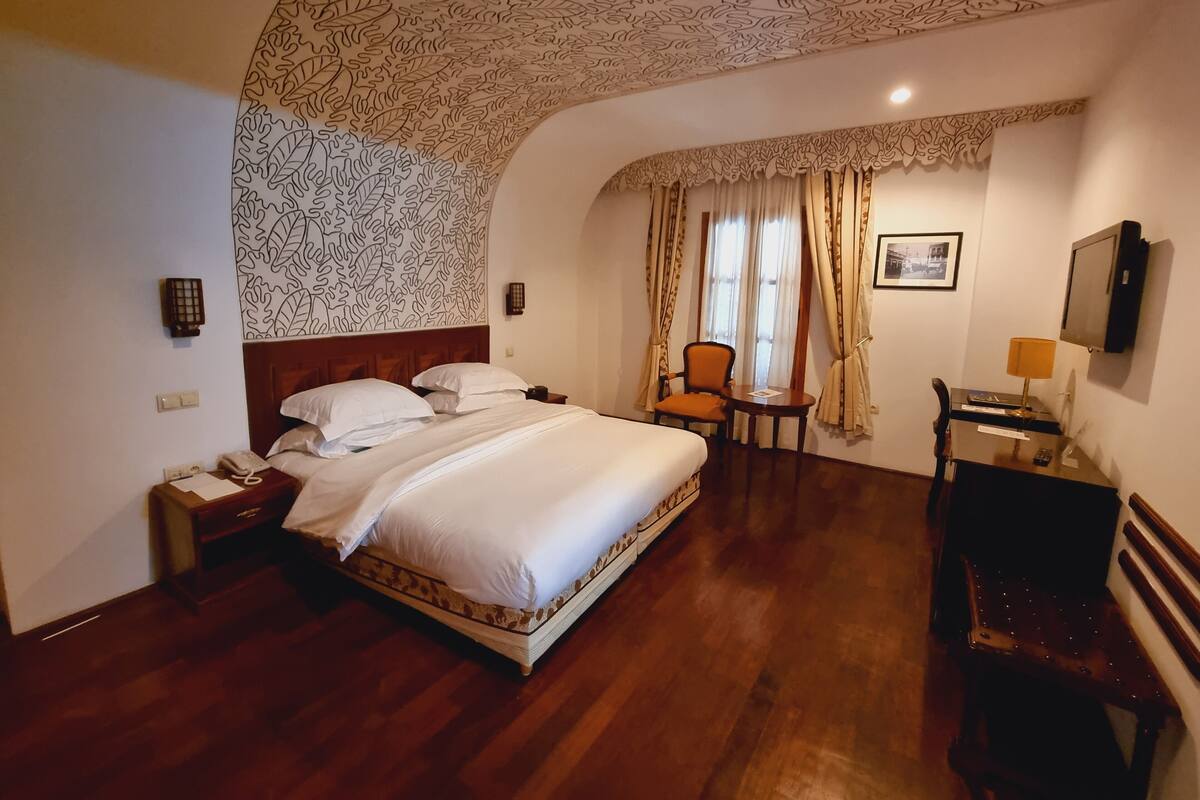 A hotel room with a large bed, a desk with a lamp, a chair, and a television on the wall.