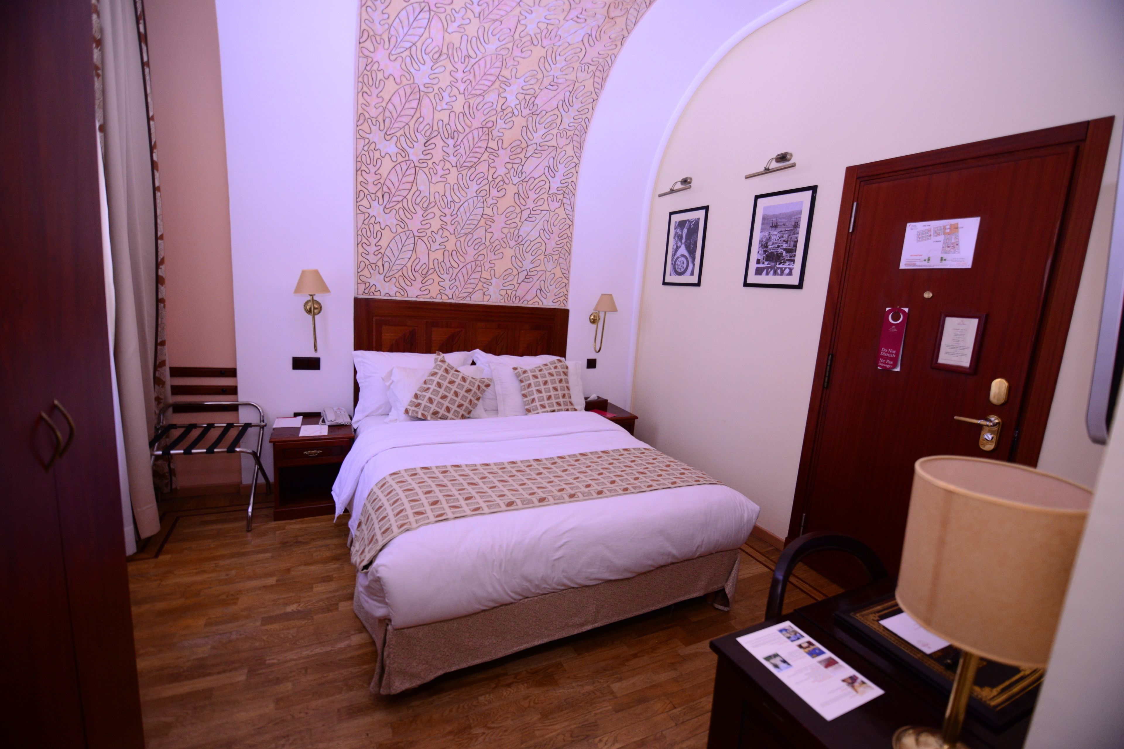 Une chambre d’hôtel avec un plancher en bois, un lit avec une literie blanche, une tête de lit en bois, deux tables de chevet avec des lampes, une armoire et une porte avec une serrure.