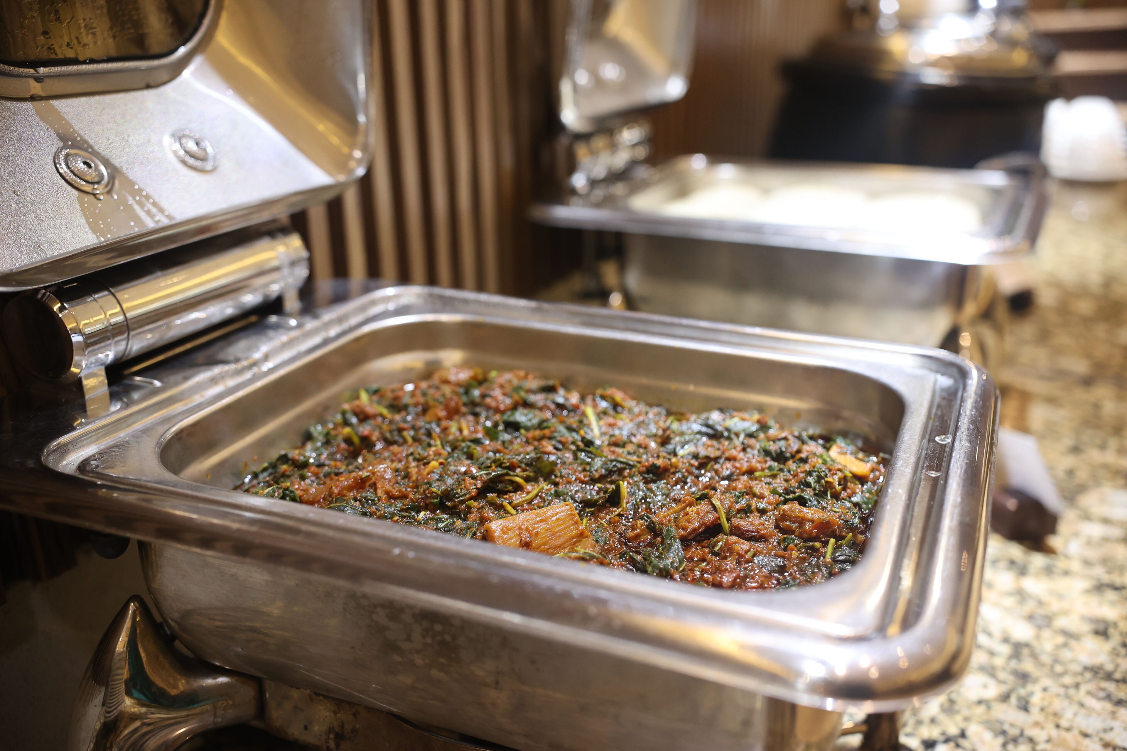 A buffet setup with a variety of dishes, including a dish with greens and possibly meat, in a stainless steel serving dish.