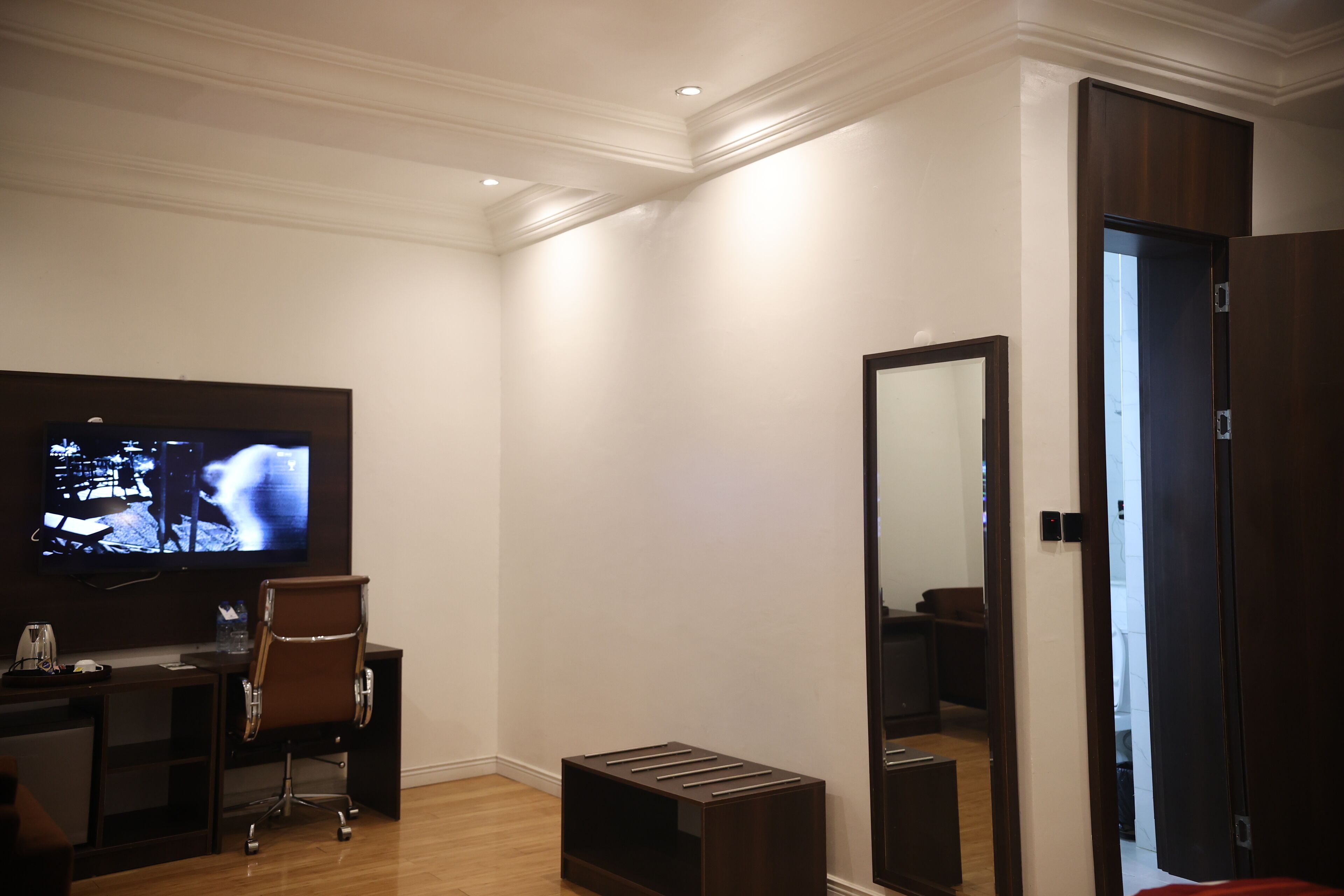 A hotel room with a flat-screen TV, a desk with a chair, a small stool, a mirror, and a wardrobe.