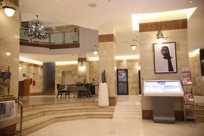 Un vestíbulo de hotel con suelo de mármol, una recepción, una escalera, una araña y un quiosco informativo digital.