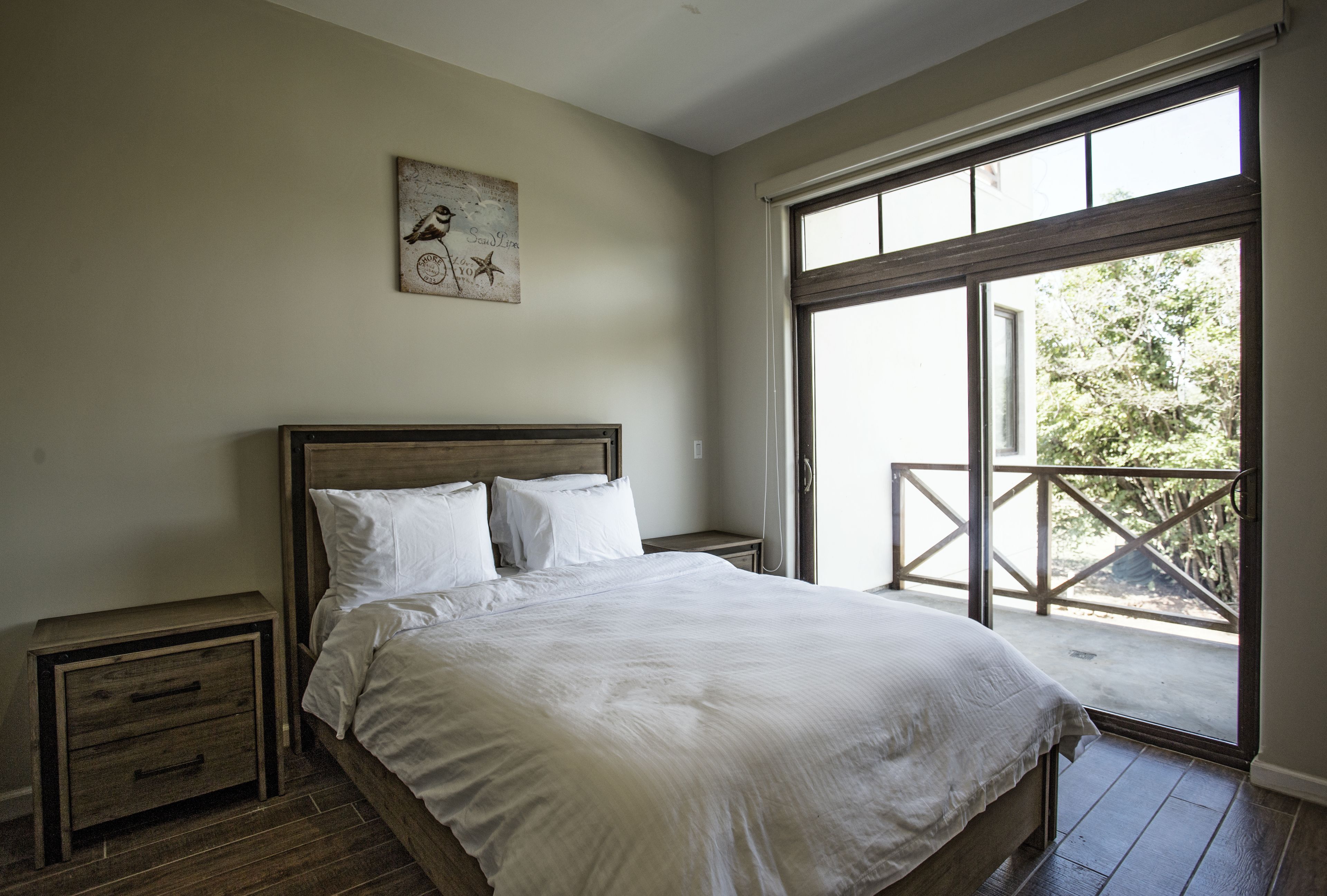 A bedroom with a large bed, wooden headboard, two bedside tables, and a view of a balcony through a sliding glass door.