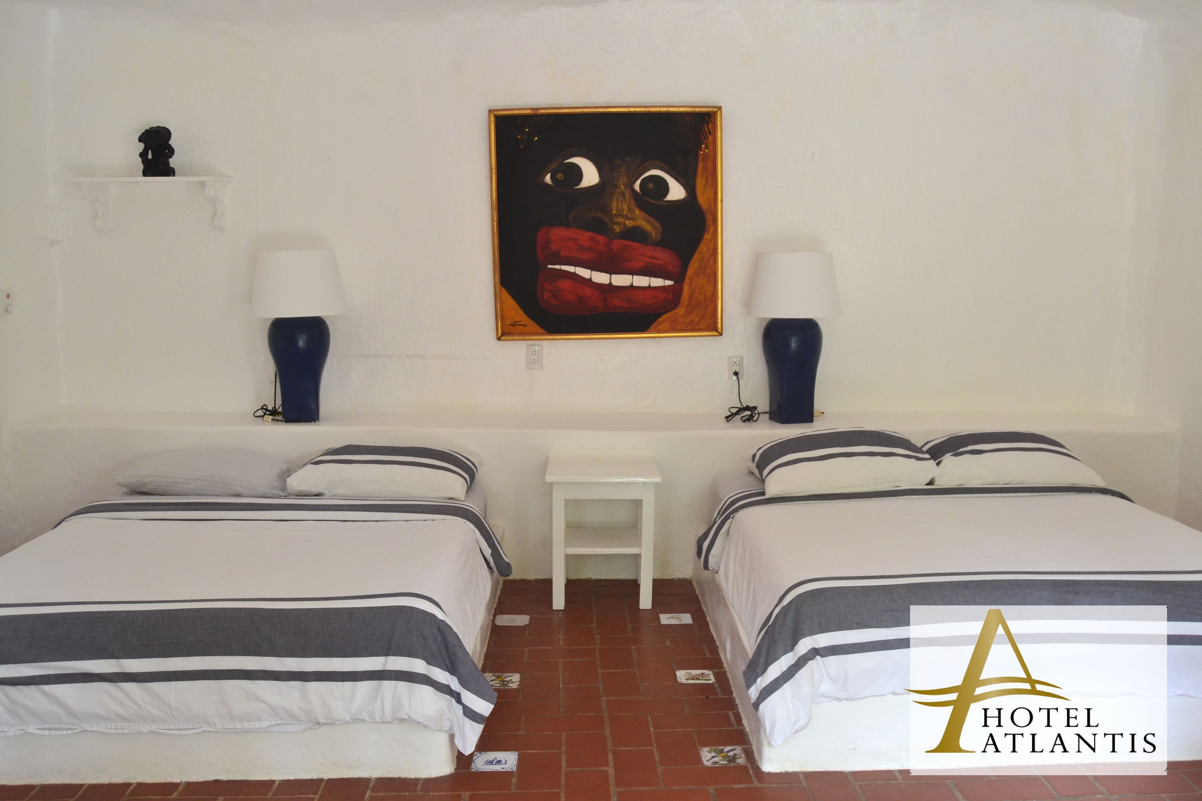 A hotel room with two beds, a small white table, and a painting on the wall.