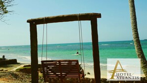 A wooden swing chair overlooking a beach with clear turquoise water and a view of the ocean. - Hotel Atlantis (Las Terrenas)