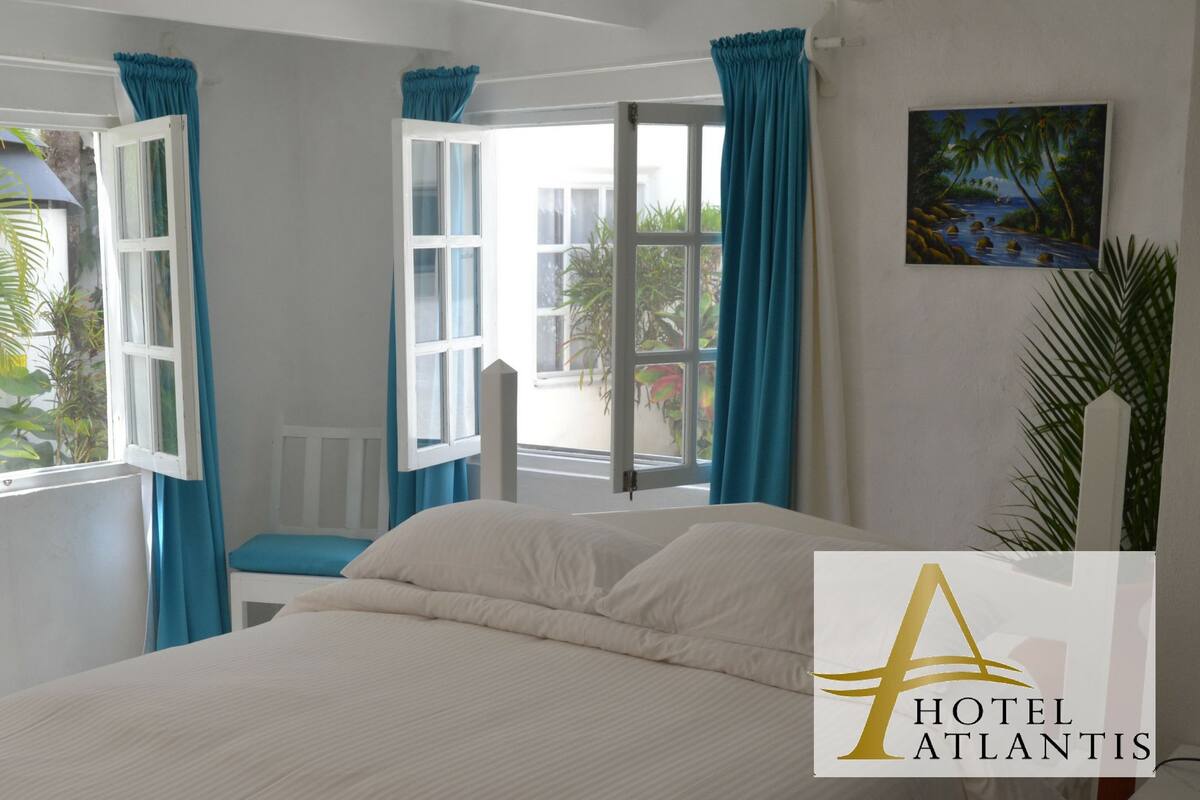 A hotel room with a bed, a desk with a lamp, and two windows with curtains.