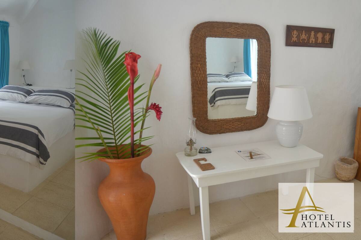 A hotel room with a bed, a white desk, a mirror, a vase with plants, and a rocking chair.