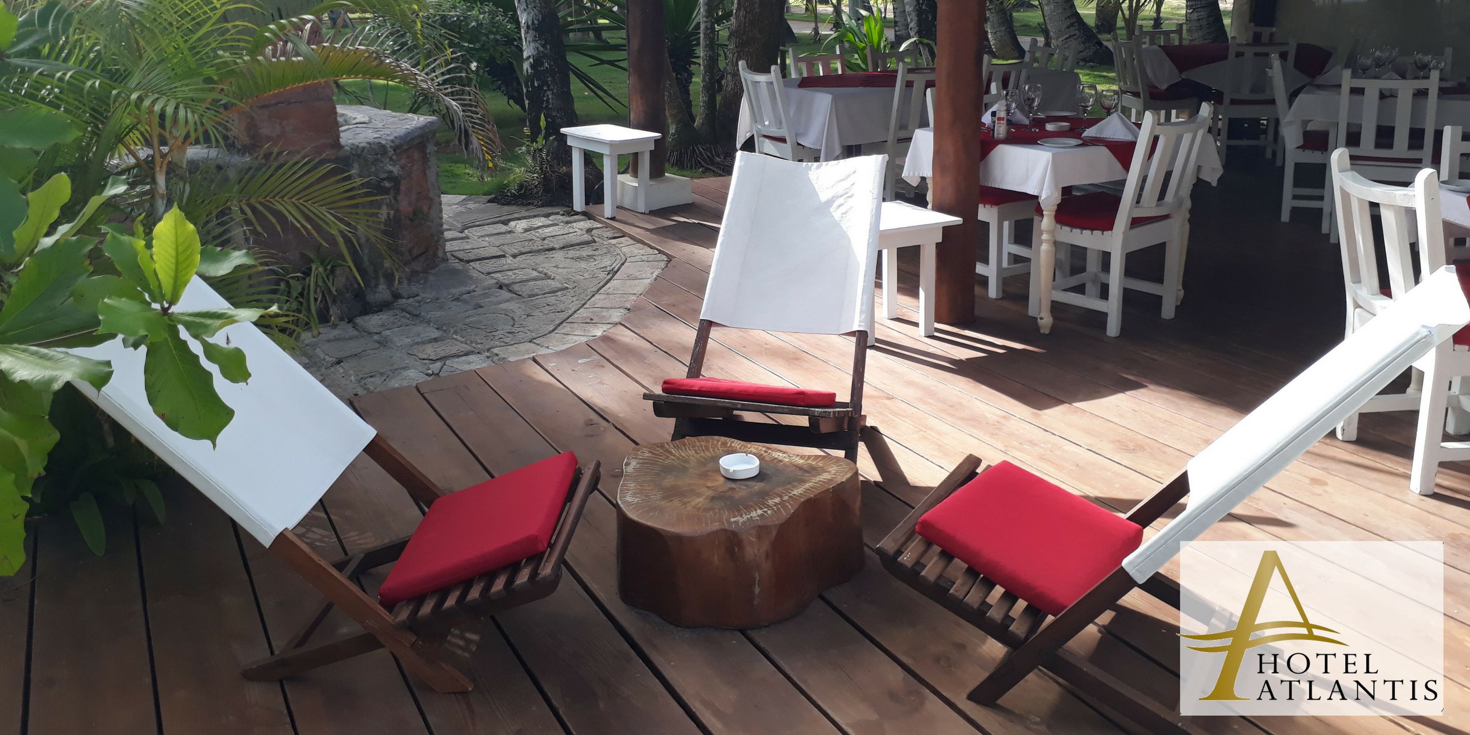 Una terraza de madera con mesas blancas y cojines rojos, rodeada de follaje y un sendero de piedra.