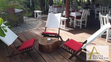 Una terraza de madera con mesas blancas y cojines rojos, rodeada de follaje y un sendero de piedra.