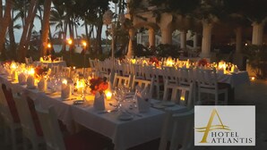 A formal outdoor dining setup with tables set for multiple guests, surrounded by palm trees and a view of the sea. - Hotel Atlantis (Las Terrenas)
