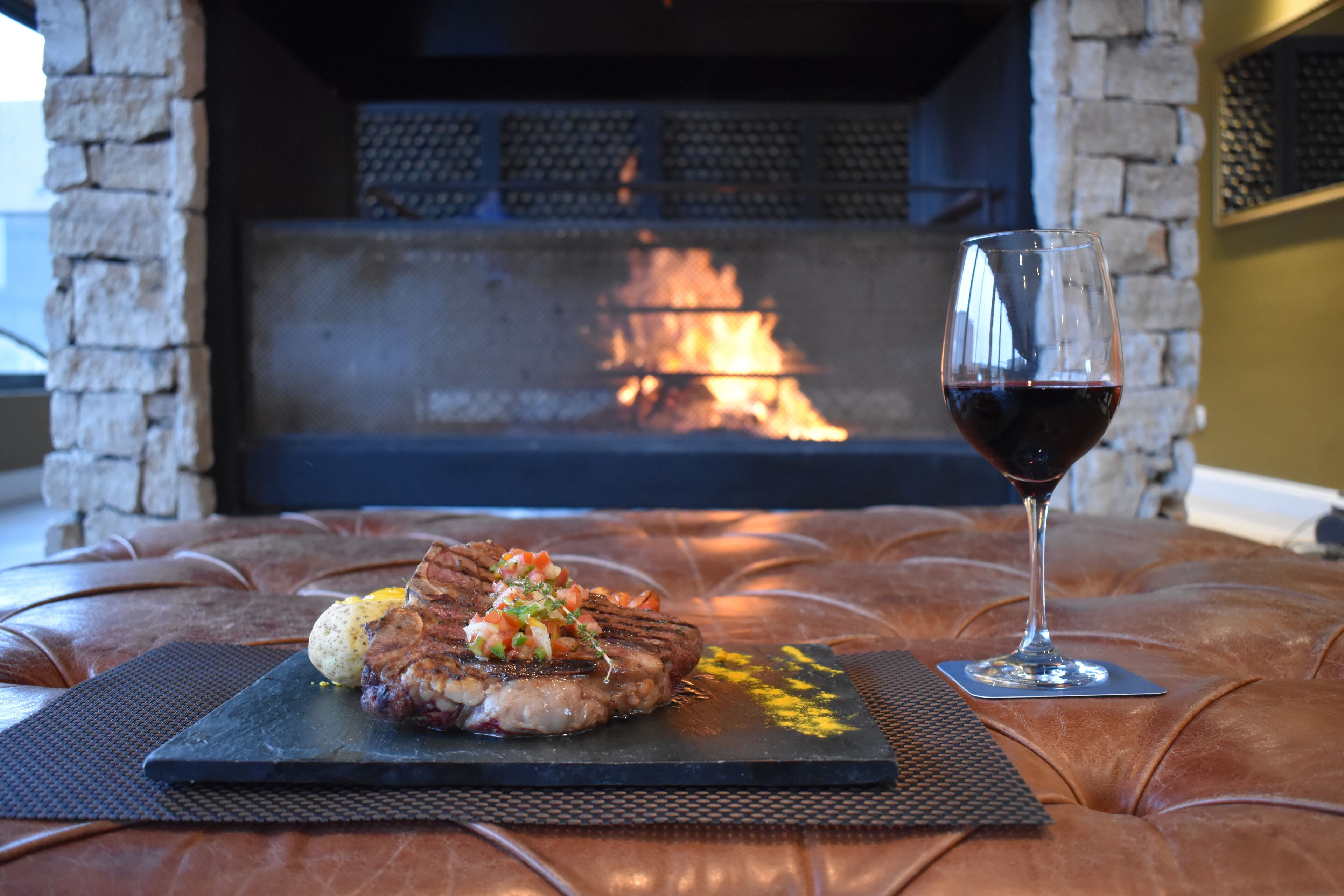 A steak with vegetables on a black slate plate, a glass of red wine, and a stone fireplace in the background.