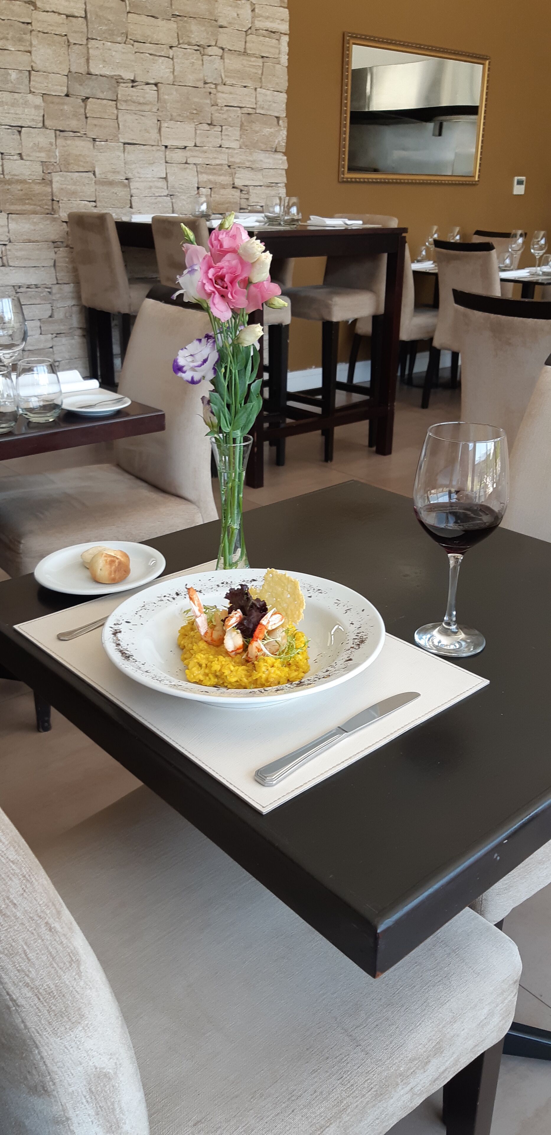 Una mesa de comedor con un plato de comida, una copa de vino tinto y un florero con flores.