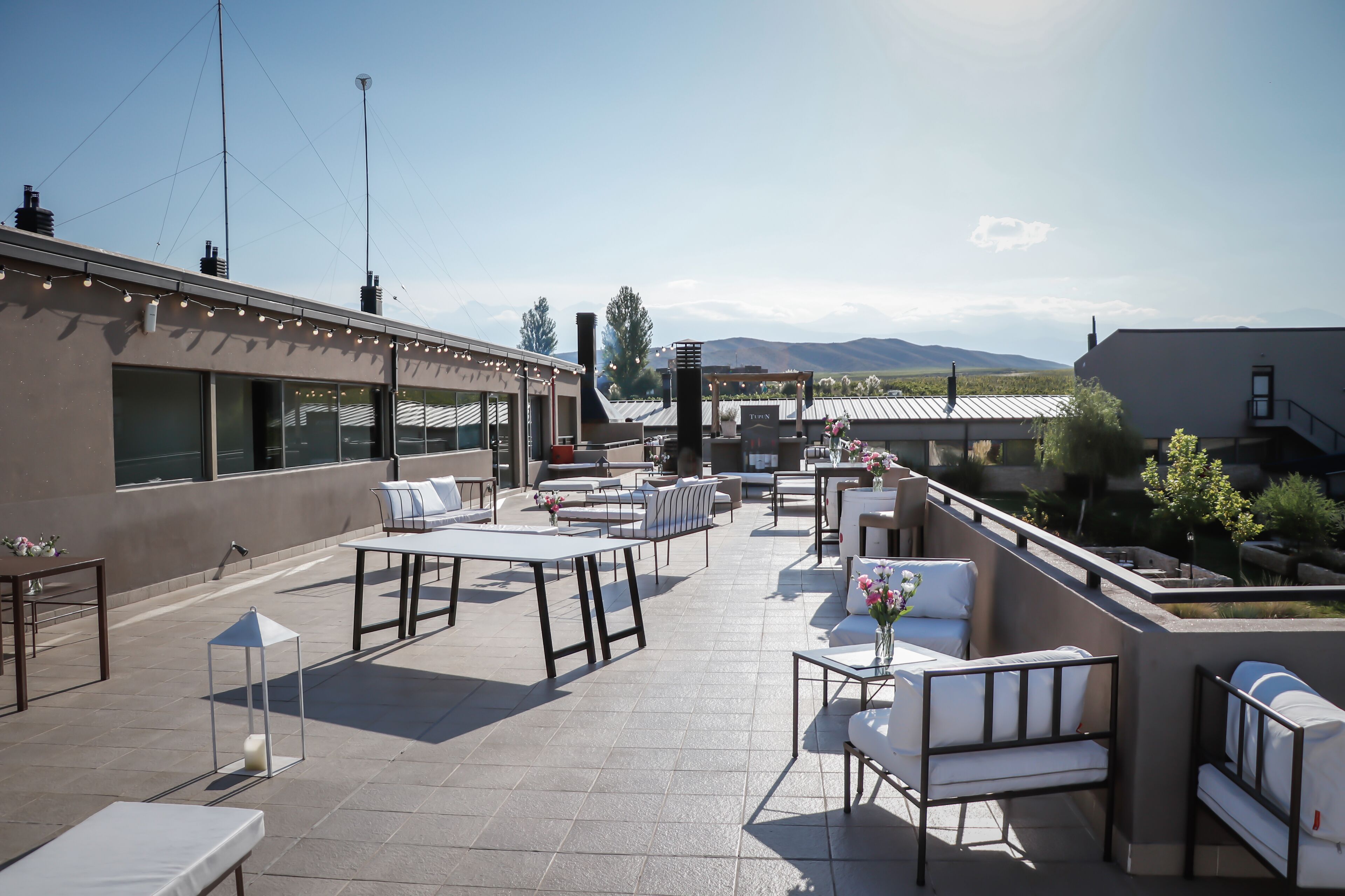 Un patio exterior con mesas y sillas, un pequeño jardín y vistas a las montañas.