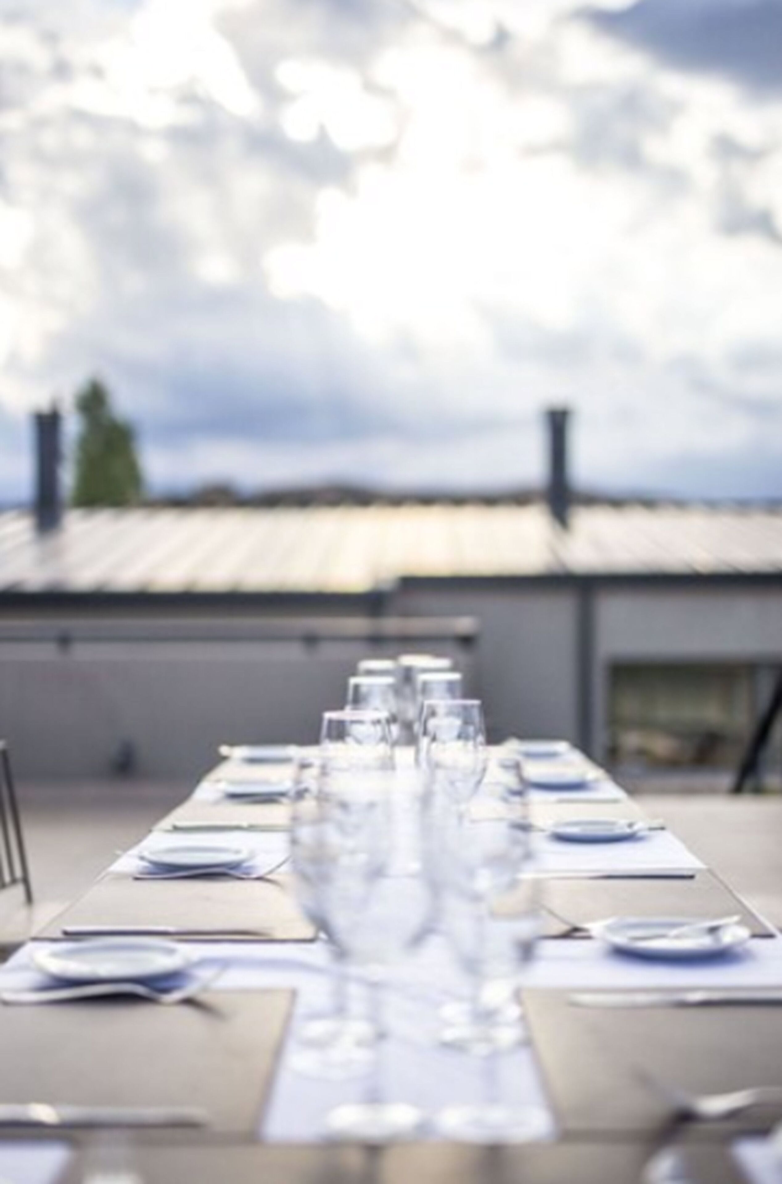 A dining table set for a meal with clear glassware and plates, positioned outdoors with a view of a building and a cloudy sky in the background.