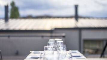 A dining table set for a meal with clear glassware and plates, positioned outdoors with a view of a building and a cloudy sky in the background.