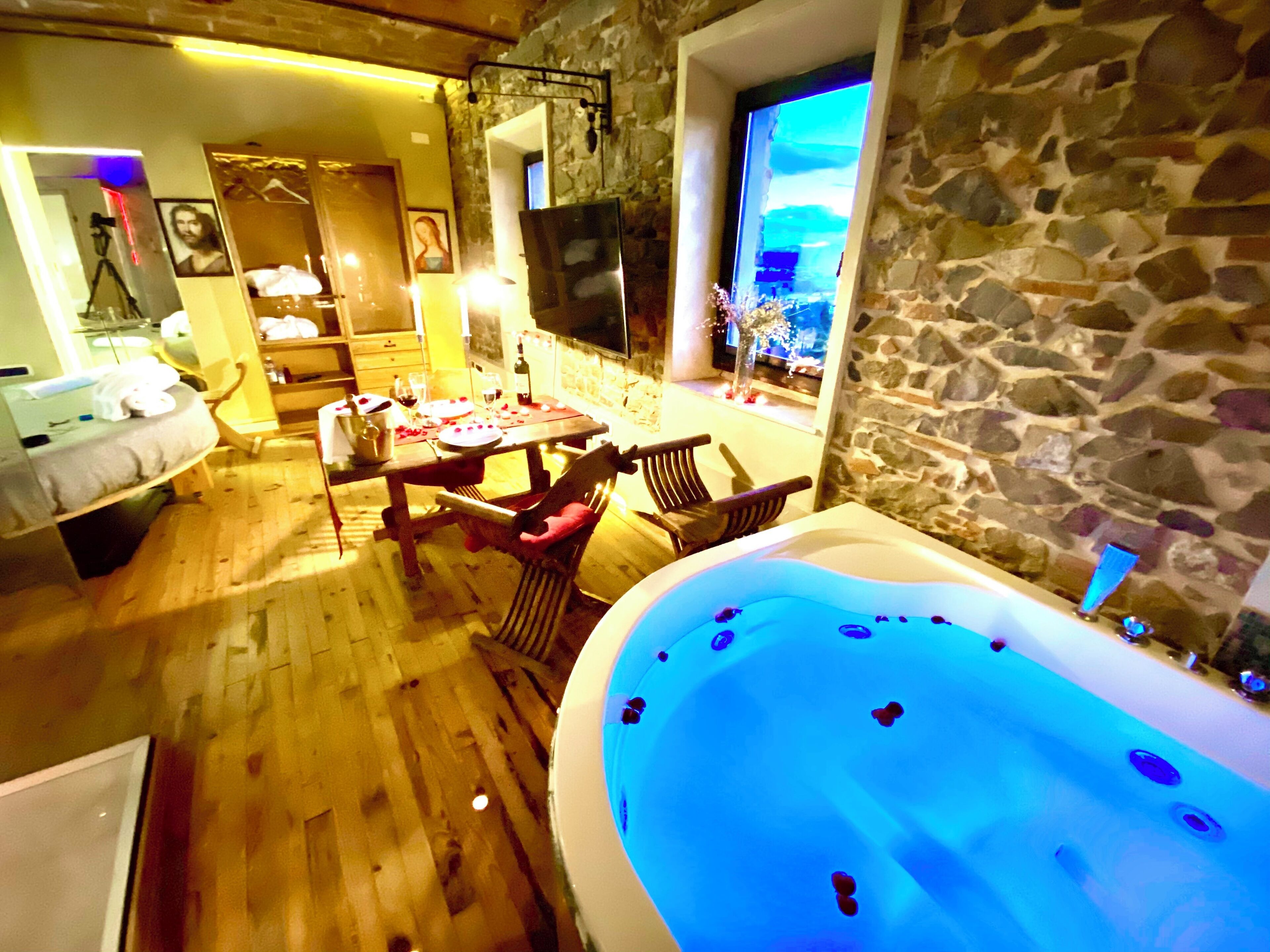 A cozy room with a stone wall, a wooden dining table, chairs, a large bathtub, and a TV mounted on the wall.