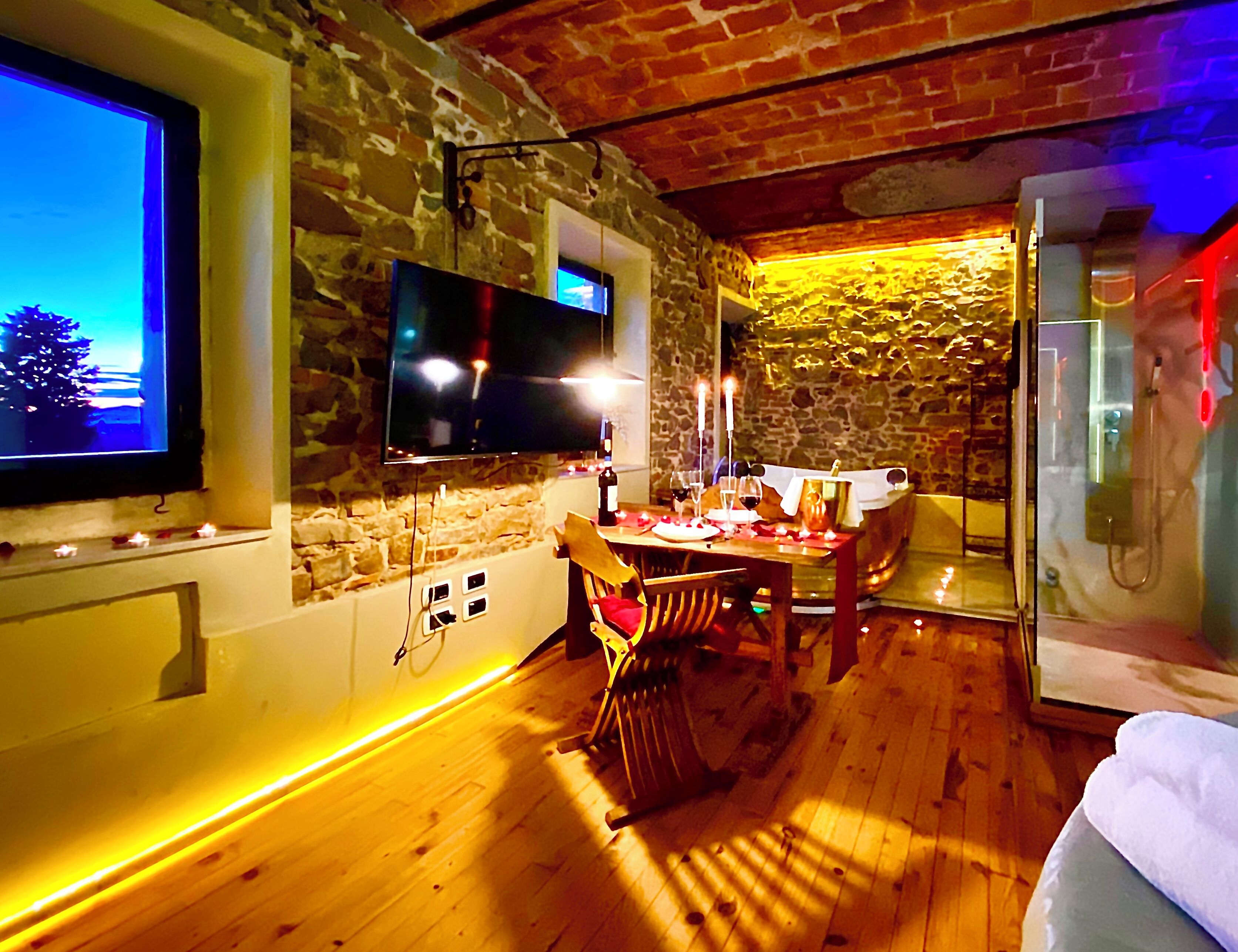 Una habitación acogedora con pared de piedra, un televisor de pantalla plana montado en la pared, una mesa de comedor de madera con velas, y una cama con edredón blanco.