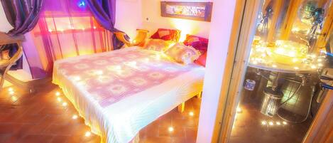 Une chambre à coucher comprenant un lit, des rideaux, une table de chevet, un miroir et une salle de bain avec un lavabo et des luminaires.
