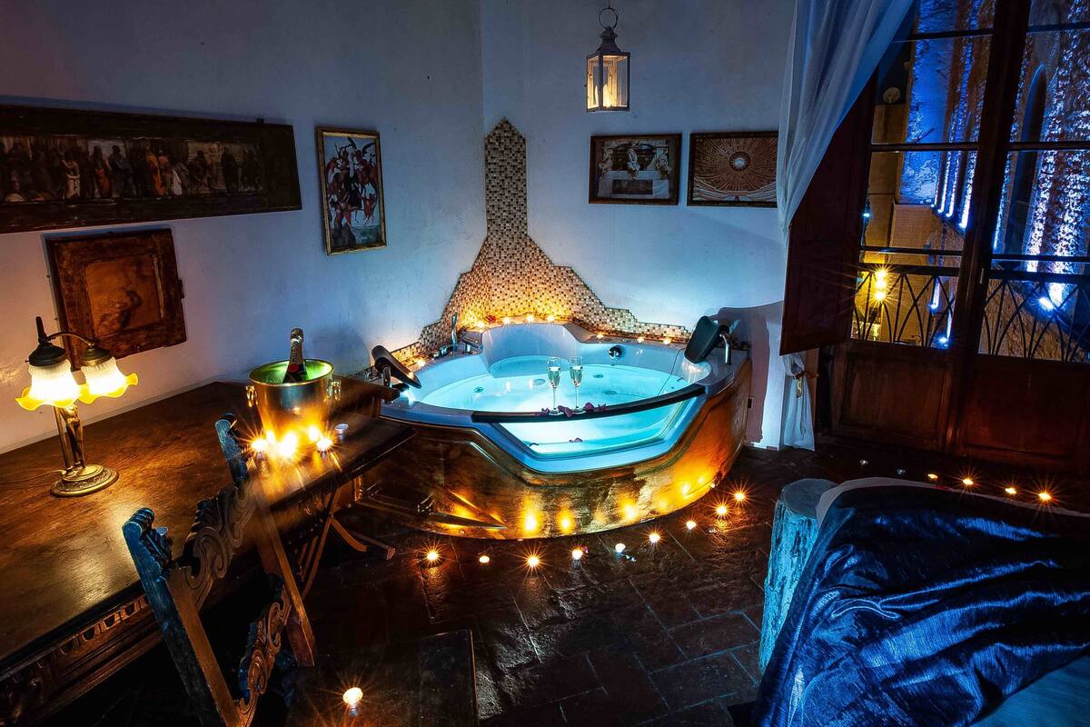 A room with a large, ornate bathtub, a wooden table with a bottle and glasses, and a window with curtains.