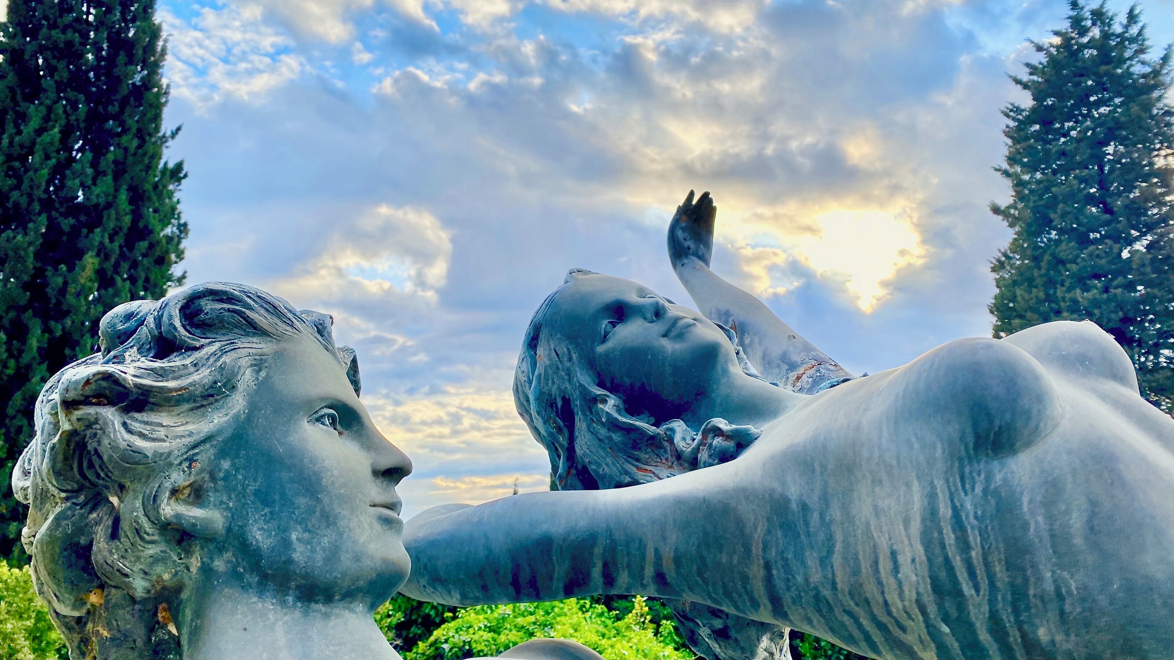 A sculpture of a reclining figure with a raised arm, set against a backdrop of trees and a partly cloudy sky.