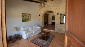 Una sala de estar con un sofá blanco, una mesita de noche con una lámpara, una mesa baja y un tapete estampado. Se observa una pared de piedra y un arco que da a otra habitación.