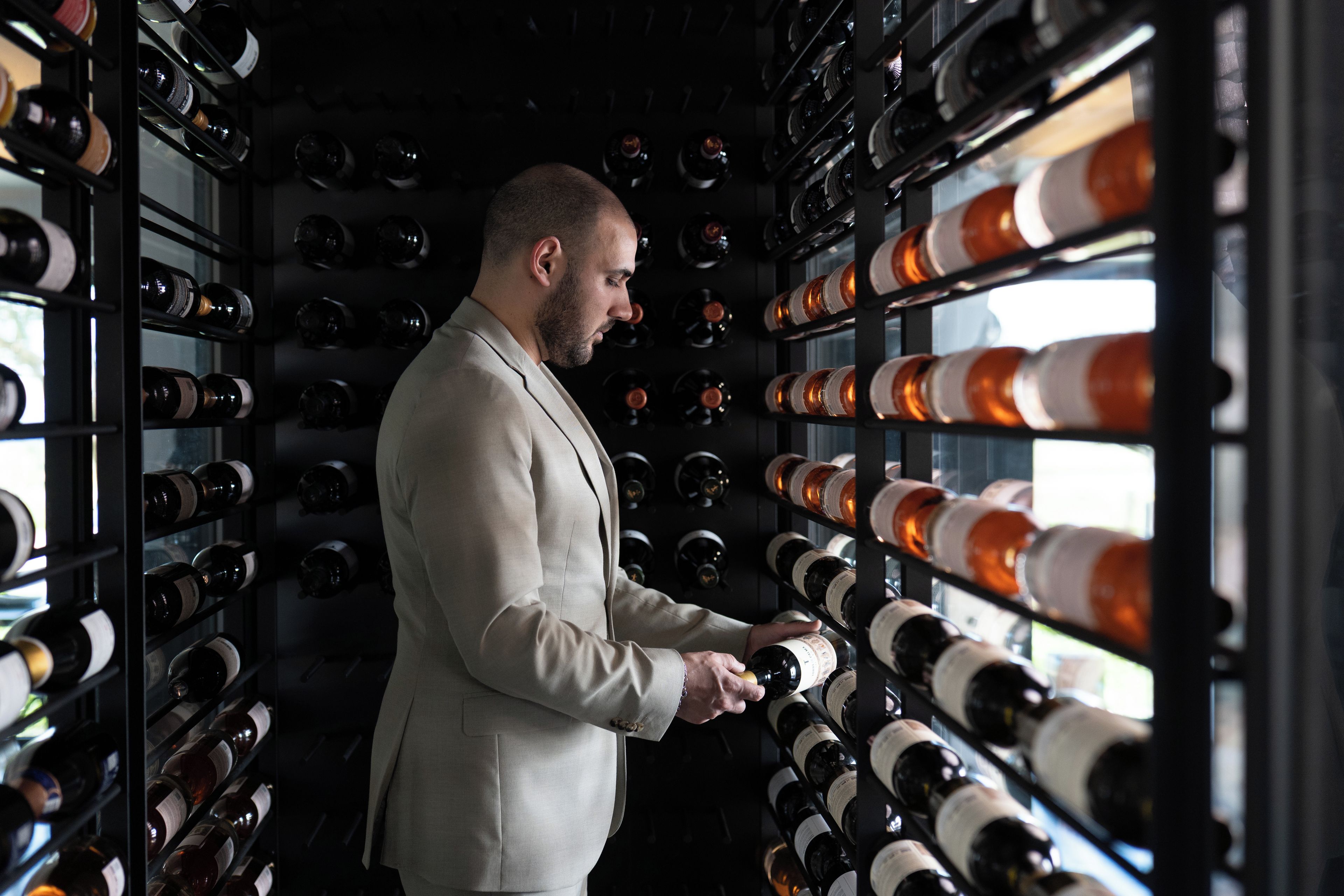 A person in a light-colored suit selecting a bottle from a wine rack in a dimly lit room.