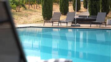 Piscina com espreguiçadeiras e um guarda-sol, cercada por sebes bem aparadas e um edifício com telhado de azulejos.