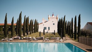 Une piscine entourée de chaises longues et de parasols, bordée de grands cyprès et d’un grand bâtiment blanc au toit rouge.