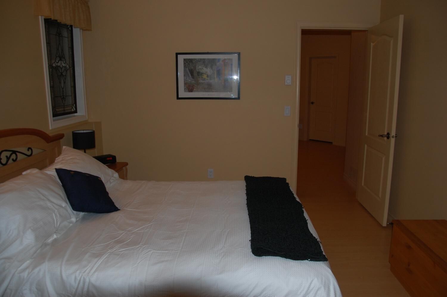 A bedroom with a bed, a nightstand, a lamp, a framed picture on the wall, and a door leading to another room.
