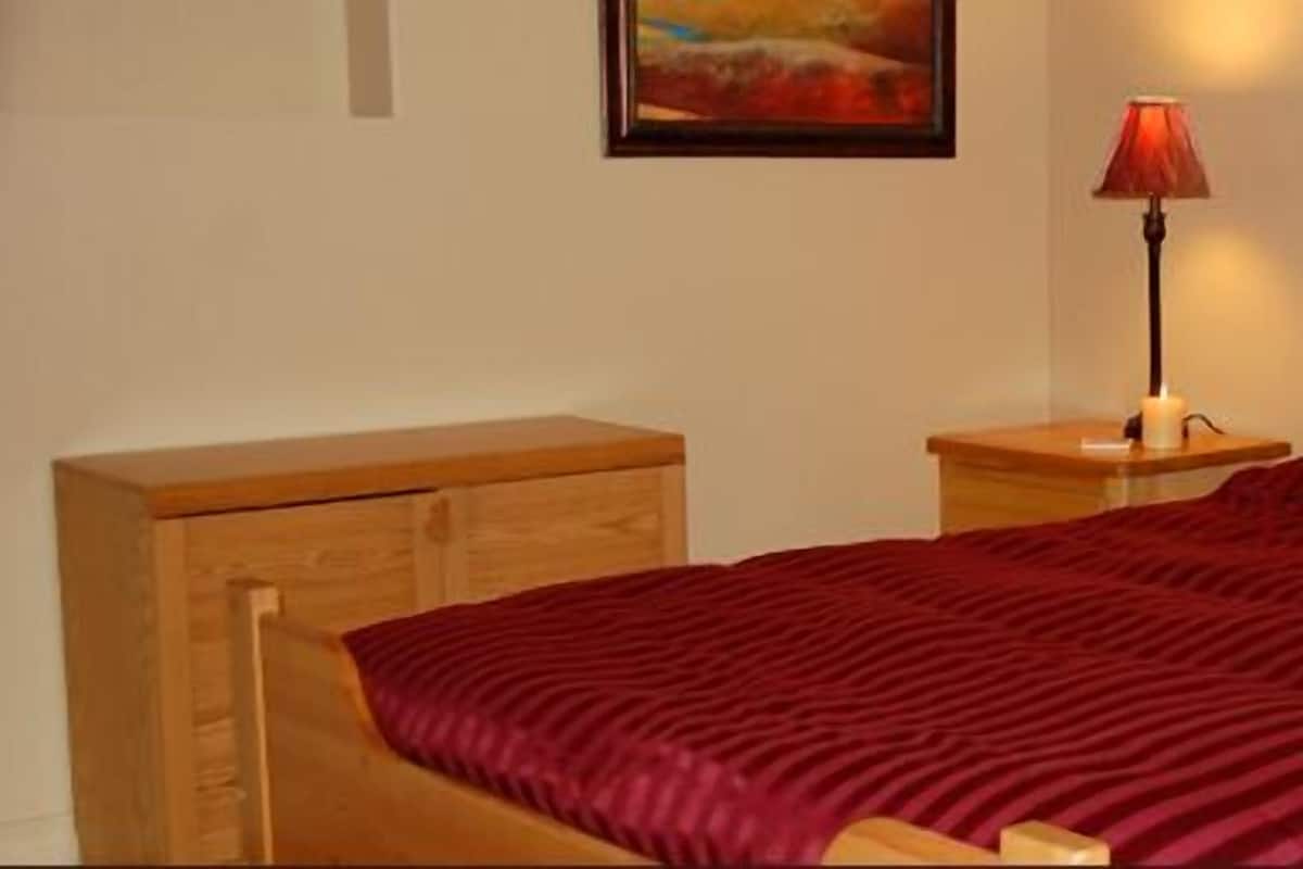 Un lit bien fait, recouvert d’une couverture rouge à rayures, une table de chevet en bois avec une lampe, et une commode en bois.