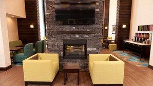 A hotel lobby with a stone fireplace, yellow armchairs, a flat-screen TV, and a coffee bar. - Hampton Inn & Suites by Hilton Regina East Gate (Regina)