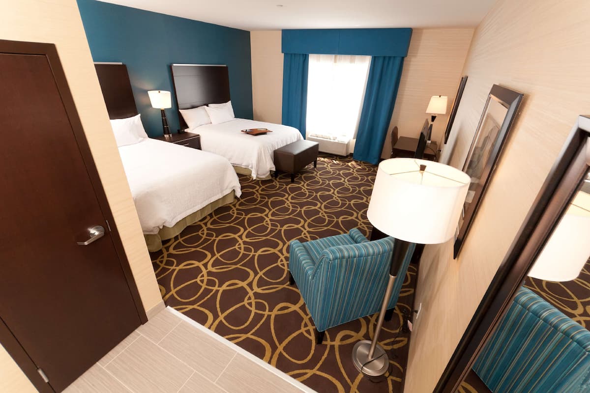 A hotel room with two beds, a desk, a chair, a lamp, and a large window with curtains.