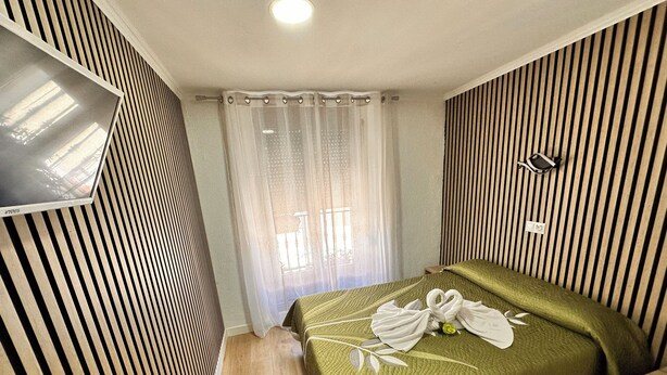 A hotel room with a bed, a TV mounted on the wall, a window with curtains, and a striped wall design. - Hostal Rober (Madrid)